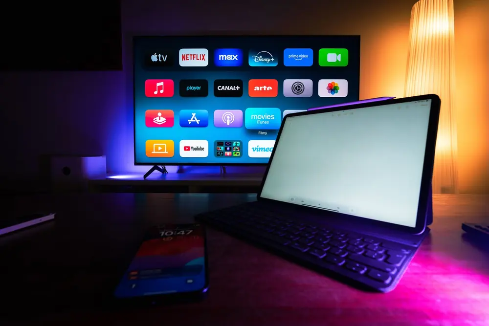 Televisão exibindo a grade de aplicativos de streaming ao lado de um notebook aberto, num ambiente com iluminação colorida