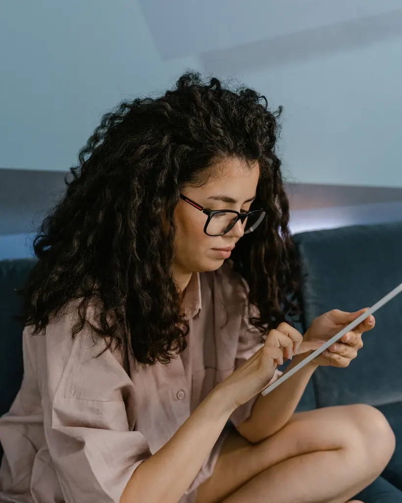 Mulher sentada de pernas cruzadas no sofá com um tablet nas mãos, escolhendo o que assistir